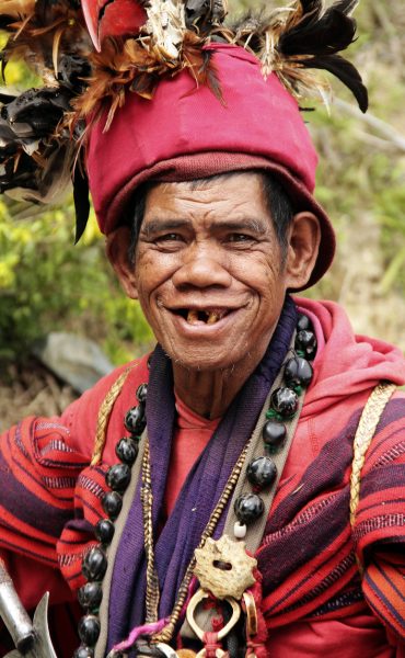 Banaue, Philippines- January 25, 2013: Head shot portrait of a native ifugao man in a traditional warrior outfit on the road to the Banaue rice terraces in the province of Ifugao, Philippines. The man is dressed in red, has feathers on his head and traditional jewellery around his knock.